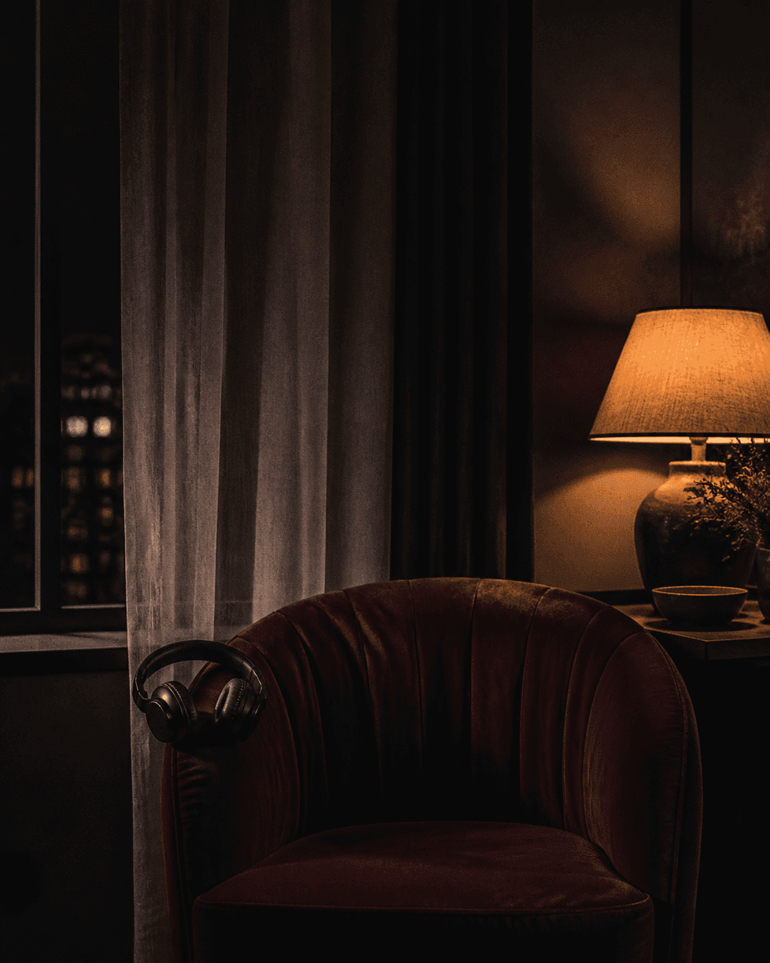 A dark room with a burgundy chair, headphones, and a lamp